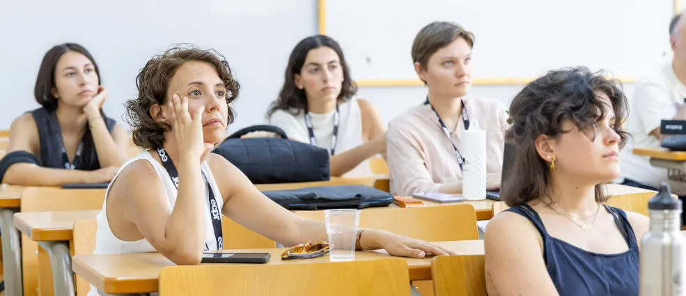 A diverse group of women attentively listening in a panel at the ECPR conference. A woman in the foreground wears a white sleeveless blouse, resting her chin on her hand.